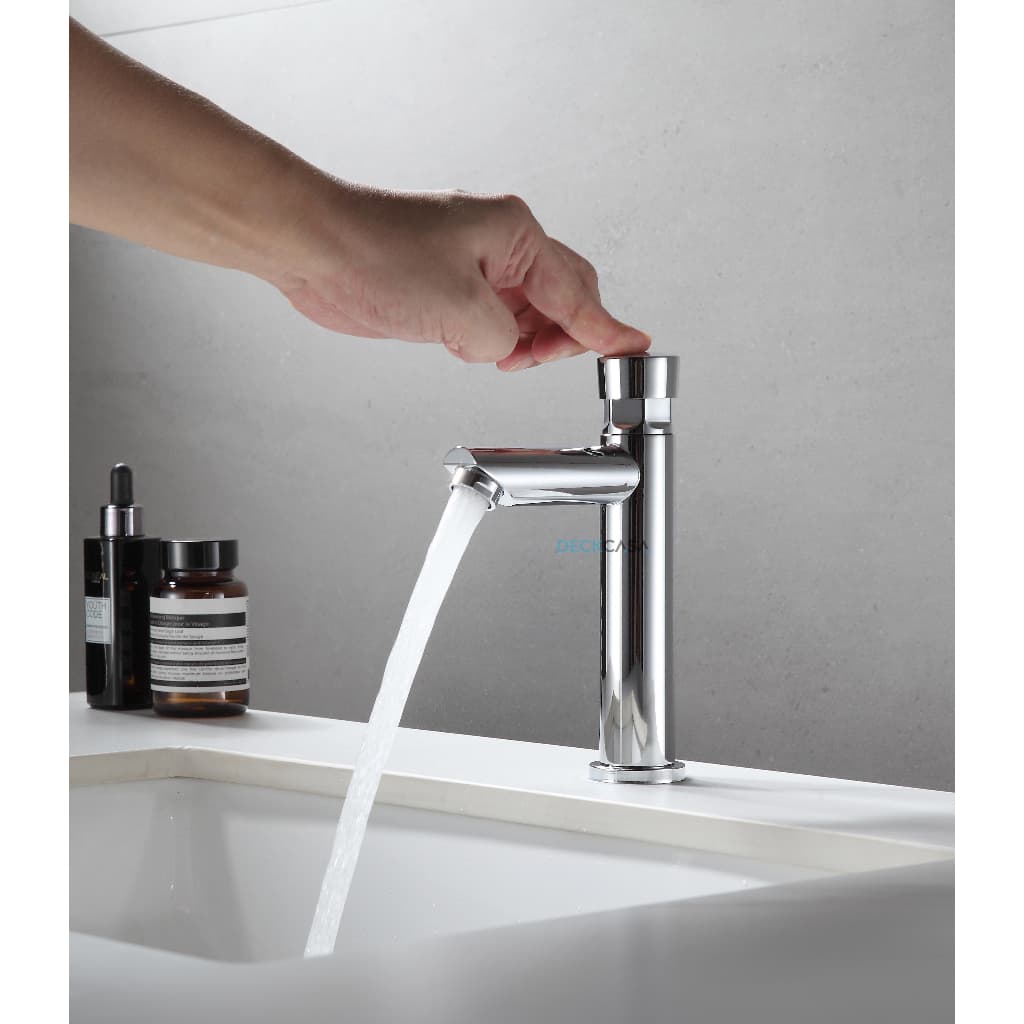 Torneira automática temporizada de metal inox para lavatório de banheiro, com água fluindo, fundo cinza e superfície branca.